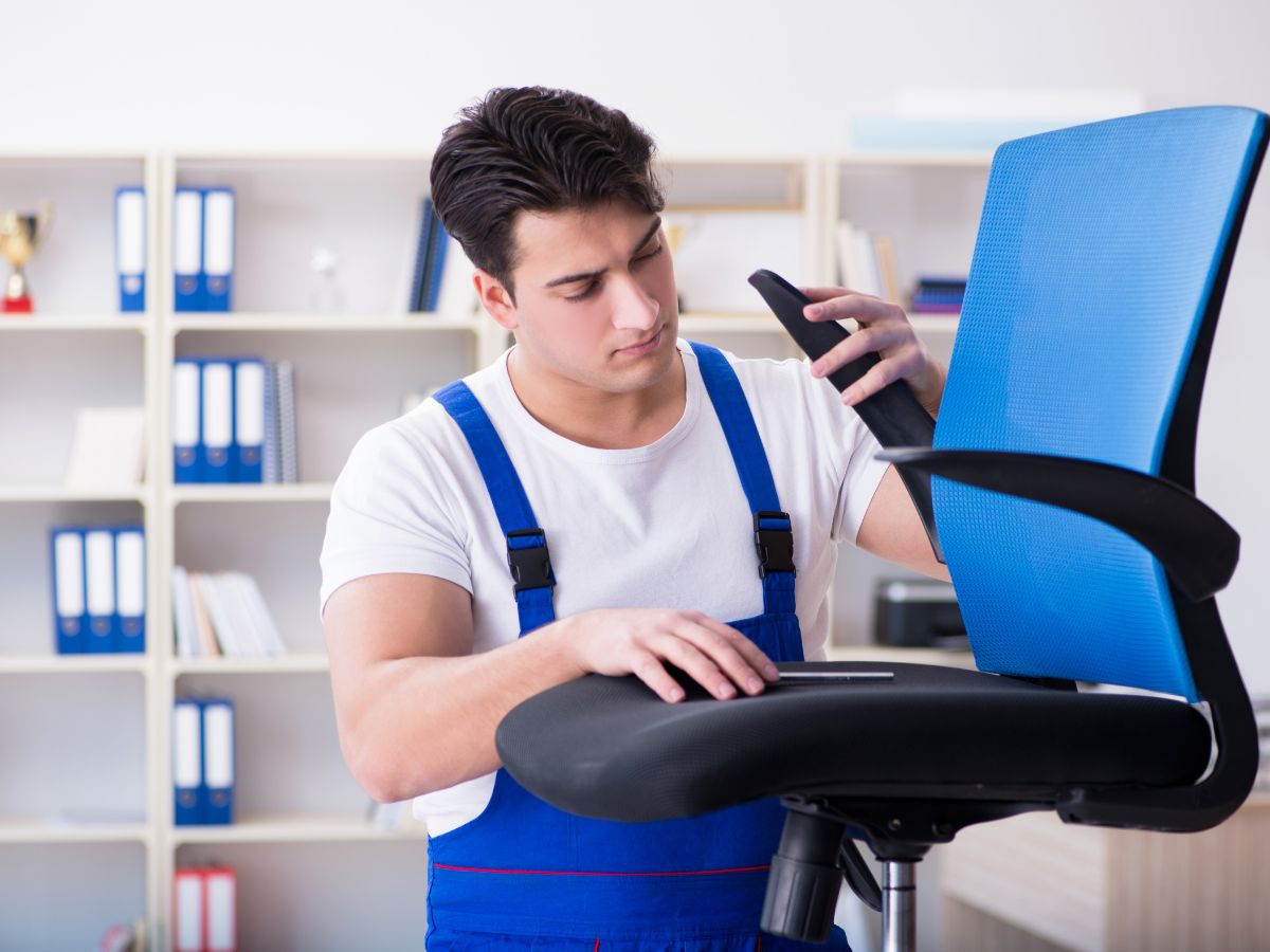 A man wearing overalls is engaged in fixing a chair, surrounded by tools in a bright workshop environment.