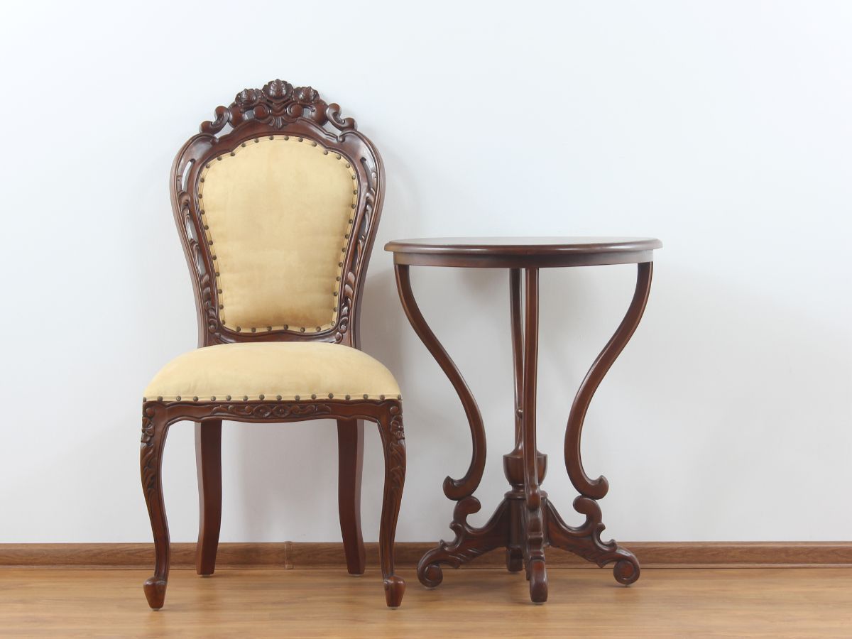 An ornate wooden chair with yellow upholstery and a matching round side table, set against a white wall.