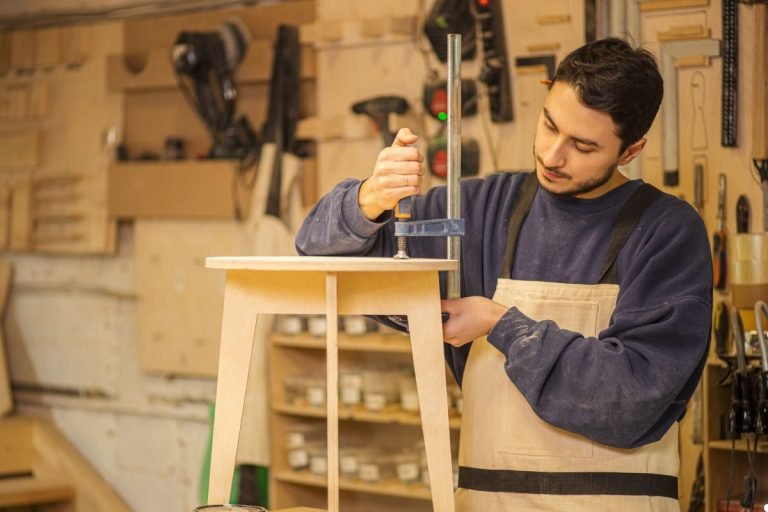 A man engaged in work at a wooden table, surrounded by tools and materials in a bright environment.