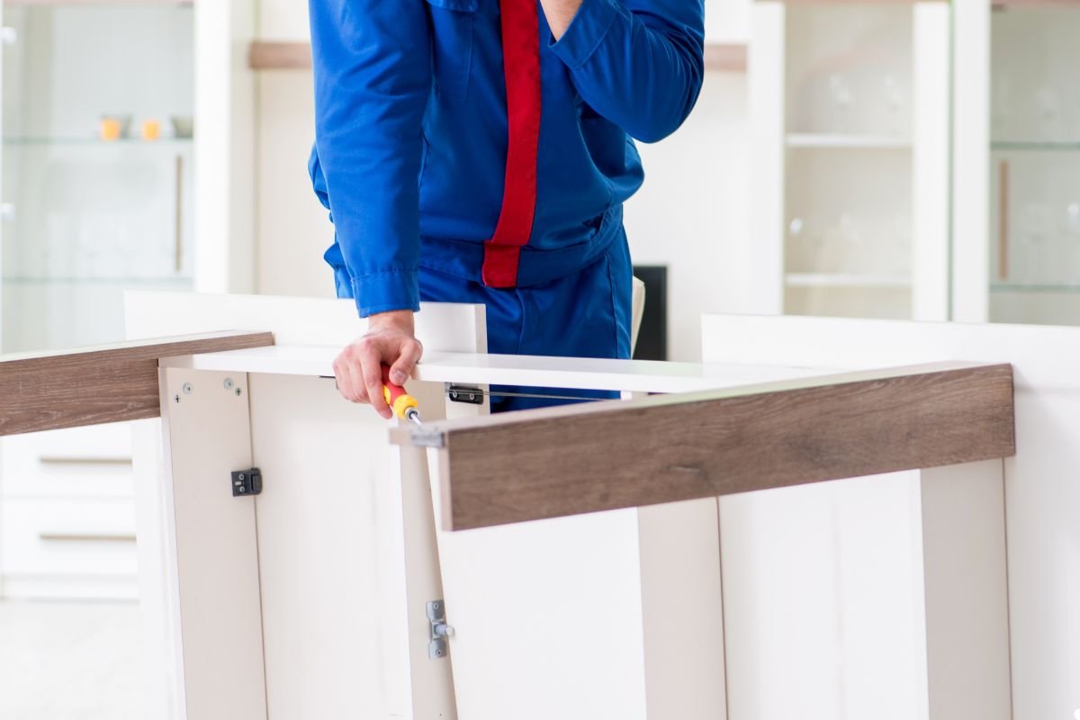 A man dressed in a blue uniform stands while holding a piece of furniture, indicating a task related to delivery or assembly.